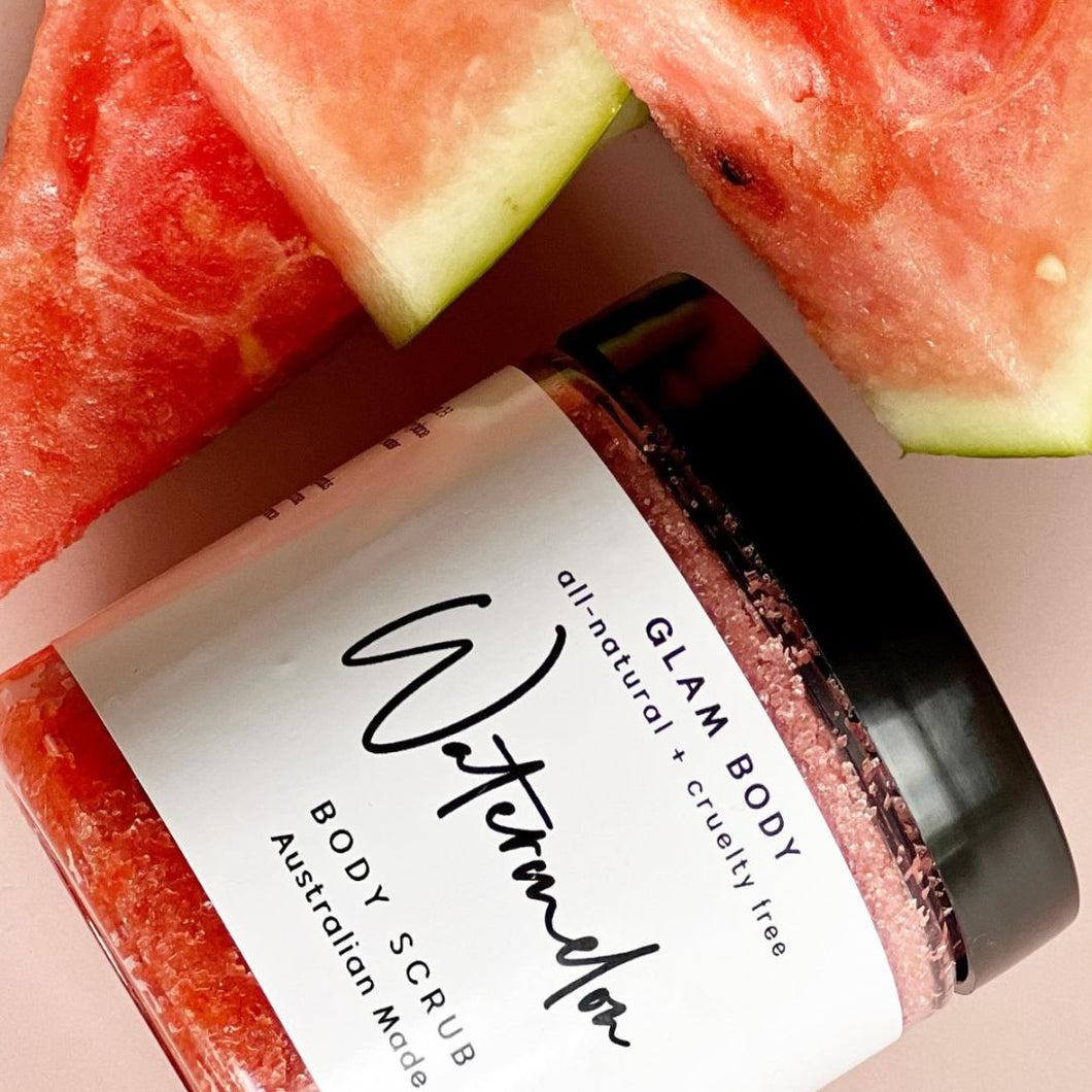 A clear Jar with Black lid containing a pink grainy scrub product surrounded by pieces of watermelon