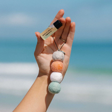 Load image into Gallery viewer, A hand holding four felt balls on a string and a small frangrance bottle in front of a out of focus beach scene
