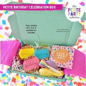 A mini aqua mailer box displaying 3 handmade cookies, 2 balloons, a party blowout and a card which says 'Have  great day!' sprinkledon a bed of shredded paper, sprinkled with confetti and enveloped in pink paper