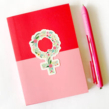 Load image into Gallery viewer, female icon symbol sticker in a floral design on a pink and red note book with pink pen
